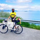 Ein Radfahrer trägt ein gelbes Trikot mit "DOWE". Im Hintergrund sind ein See und Berge zu sehen.