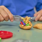 Ein Kind formt bunte Knete mit Werkzeugen. Es trägt ein blaues Oberteil. Farbiges Zubehör liegt auf dem Tisch.