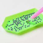 Ein grünes Tablett mit lila Perlen und einem rosa Werkzeug zum Aufnehmen der Perlen.