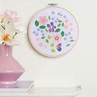 Ein Blumenmotiv in einem Stickrahmen hängt an der Wand. Daneben steht eine rosa Vase mit Blumen auf einem Regal.