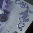Text: "..ES". Geprägte, violette Ornamente und Rosen auf einer grauen Fläche.