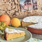 Text "PALMA" auf einer Postkarte mit Palmen. Ein Mandelkuchen, eine Orange, eine Zitrone und gelbe Blumen.