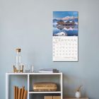 Kalenderblatt mit "JANUAR 2023", Monatsübersicht. Darunter Holzhäuser im Schnee vor Bergen. Schlichtes Regal mit Karaffe.