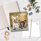 Kalender mit "Hunde 2027" und zwei Welpen. Daneben ein offenes Notizbuch, Stift, Postkarte, Briefmarken und Eukalyptuszweig.