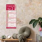 Kalender "TIMER for 2 2027" an einer strukturierten Wand. Darunter Kissen, Pflanze, Uhr auf Tisch.