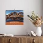 2027 HÜTTENZauber DIE ALPEN. Ein Kalender hängt an einer Wand, darunter ein Sideboard mit Büchern und einer Vase.