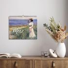 Text: „Am Meer“ und „2024“. Wandkalender mit einem Gemälde einer Frau am Strand. Darunter ein Tisch mit Büchern und einer Vase.