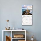 "Juli 2027" steht auf einem Wandkalender mit einem Bild eines Leuchtturms auf einer Klippe. Darunter ein Regal mit Deko.