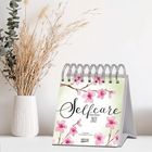 Kalender mit „Selfcare Katja Reiter 2021“. Illustration von rosa Blüten. Steht neben einer Vase mit getrockneten Gräsern.