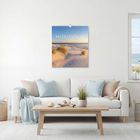 Text "MEERESSTILLE 2024". Wohnzimmer mit Sofa, helle Kissen, Holz-Couchtisch, Dünenbild an der Wand.