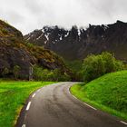 Eine kurvige Straße führt durch grüne Hügel, gesäumt von Bäumen, vor steilen dunklen Bergen und bewölktem Himmel.