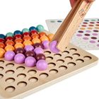 Bunte Holzkugeln auf einem Holzbrett mit Löchern; eine mit Holzpinzette gehalten. Im Hintergrund eine Farbvorlage.
