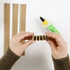 Text: "BASTELKLEBER", "HOBBY GLUE". Zwei Hände falten einen Papierstreifen, daneben Kleber und drei Kartonstreifen.
