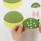 Hände halten ein grünes Papierstück mit weißen Quadraten. Größere grüne und gelbe Kreise im Hintergrund.