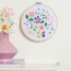 Stickerei mit bunten Blumen im Stickrahmen. Daneben eine rosa Vase mit Blumen auf einem weißen Tisch.