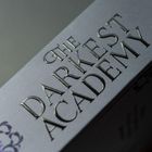 Text: "THE DARKEST ACADEMY". Dunkler Hintergrund, erhabene Schrift in stilvoller Typografie.