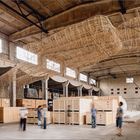 Große Halle mit Menschen, Holzkisten und einem großen hängenden Objekt aus geflochtenem Material an der Decke.