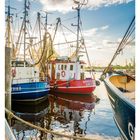 Beschriftung unten: "APRIL", Wochentage und Daten. Drei Fischerboote im Hafen, bunte Farben, ruhiges Wasser, blauer Himmel.