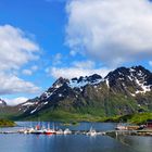 Hafen mit Booten vor schroffen Bergen und blauem Himmel, teils mit Wolken. Eine kleine Hütte am Ufer.