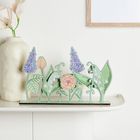 Eine beige Vase mit dezenten Blumen steht auf einem Regal. Daneben dekorative florale Illustrationen in Pastellfarben.
