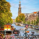 Drei Fahrräder auf einer Brücke über einem Kanal, im Hintergrund eine Kirche mit hohem Turm, umgeben von Bäumen.