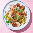 Es gibt keinen Text auf diesem Bild. Ein weißer Teller mit Reisnudeln, Tofu, Gurkenscheiben und Koriander auf rosa Hintergrund.