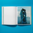 Eine aufgeschlagene Buchseite mit einem Bild einer alten Kamera am Ufer, in blauen Stoff gehüllt, vor Wasser und Bergen.