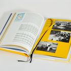 Ein aufgeschlagenes Buch mit Tabellen und Schwarzweißfotos auf gelbem Hintergrund. Ein Lesezeichen liegt im Buch.