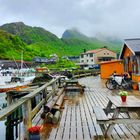 Ein Holzsteg mit Bänken, Blumen, Fahrrädern und Booten. Im Hintergrund sind grüne Berge und ein grauer Himmel zu sehen.