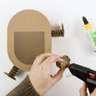 Text: "BASTEL". Eine Hand hält Pappstück und Heißklebepistole. Daneben liegt ein Bienenstock aus Pappe mit gefalteten Armen.