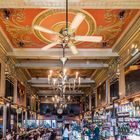 "EXIT"-Schilder an den Wänden. Reich verzierte Decke, Kronleuchter, Bar, Menschen in einem Kaffeehaus.