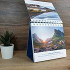 Es zeigt den Text: "Es ist eins der großen Geschenke der Stille, dass sie uns darauf aufmerksam macht, wo wir uns befinden." Ein Kalender steht auf einem Tisch, daneben eine kleine Pflanze und einige Steine.