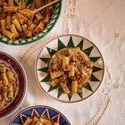 Verschiedene bunte Schüsseln und Teller mit kurzen Rigatoni-Nudeln und Soße auf einem bestickten Tischtuch.