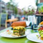Ein Teller mit einem hohen veganen Burger aus Gemüse und Sprossen steht im Freien auf einem grünen Tisch.