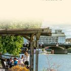 Text: "PRENZLAUER BERG FRIEDRICHSHAIN KREUZBERG". Eine Flusspromenade mit Menschen auf Holzbänken, Pflanzen im Vordergrund.