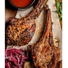 Zwei gegrillte Lammkoteletts auf einem Holzbrett, daneben rote Zwiebeln, Pilze und Schüssel mit roter Soße.