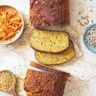Hellbraunes Brot mit zwei Scheiben, umgeben von Schalen mit Karottenstreifen, Sonnenblumenkernen und Leinsamen.