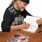 Ein Mann mit Bart und schwarzem Tuch signiert ein Buch. Mehrere gleiche Bücher liegen auf einem Tisch davor.