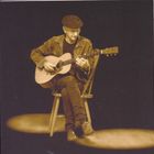 Ein Mann mit Hut spielt Gitarre, sitzt auf einem Stuhl, gedämpftes Licht, trägt Jeansjacke und -hose.