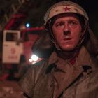 Der Text zeigt "6967" auf einem Feuerwehrauto. Ein Feuerwehrmann, schmutzig und erschöpft, steht im Vordergrund.