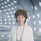 Eine ältere Frau mit lockigem Haar in weißer Kleidung steht vor einem futuristischen Hintergrund mit runden Fenstern.