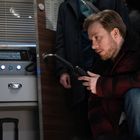 Ein Mann in roter Jacke kniet, spricht in ein Walkie-Talkie. Links ein Kasten mit roten und blauen Knöpfen.