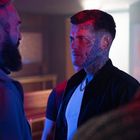Zwei Männer stehen sich gegenüber in einem beleuchteten Raum, einer hat Gesichtstattoos, der andere einen Bart.