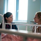 Zwei Frauen unterhalten sich in einem Krankenzimmer. Eine trägt ein Stethoskop. Im Vordergrund ein Krankenhausbett.
