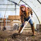 Es sind keine Texte sichtbar. Eine Frau mit rotem Haar gräbt Erde mit einer Schaufel in einem Gewächshaus um.