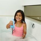 Ein lachendes Mädchen in einer Badewanne voller Schaum hält ein blaues Wasserspielzeug. Sie trägt ein rosa Oberteil.