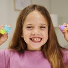 Ein lachendes Mädchen in lila Oberteil hält bunte Spielzeuge in den Händen, eins blau-grün, das andere lila-gelb.