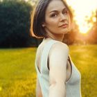 Eine Frau blickt über die Schulter zurück und hält eine Hand, im Hintergrund eine Wiese mit Sonnenuntergang.