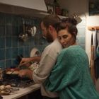 Ein Paar steht in einer Küche; der Mann kocht, während die Frau ihn liebevoll umarmt. Die Atmosphäre wirkt gemütlich.