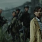 Kein sichtbarer Text. Ein Mädchen in gelber Jacke blickt nachdenklich. Im Hintergrund unscharfe Soldaten in Tarnanzügen.